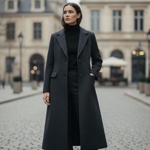 May include: A long, dark gray coat with a classic design. The coat features a notched collar, front pockets, and a long, flowing silhouette. The person in the image is wearing a black turtleneck and black pants. The coat is a stylish outerwear option.