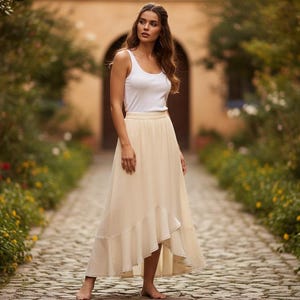 May include: A woman wearing a white tank top and a cream-colored, flowing skirt stands on a cobblestone path. The skirt has a layered, asymmetrical hemline. The background features lush greenery and a stone archway, creating a natural, elegant setting.