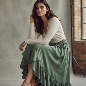 May include: A woman wearing a cream-colored long-sleeve top and a sage green skirt with a ruffled hem. The skirt is a flowing, midi-length design. The background is a neutral gray with a brick wall and window visible.