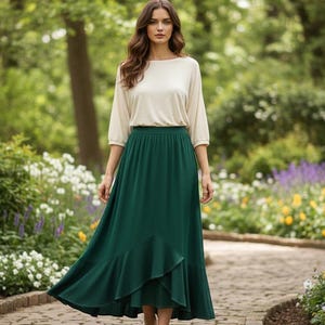 May include: A woman wearing a cream-colored top with three-quarter sleeves and a long, flowing emerald green skirt. The skirt has a ruffled hem. She is standing on a stone path in a garden with flowers and trees.