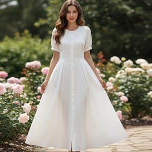 May include: A white, button-down dress with short sleeves and a full skirt. The dress is worn in a garden setting, surrounded by pink and white roses. The dress has a classic, elegant design.