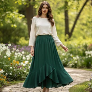 May include: A woman wearing a cream-colored long-sleeve top and a long, flowing emerald green skirt. The skirt has a ruffled hem. She is walking on a stone path in a garden with flowers and trees.