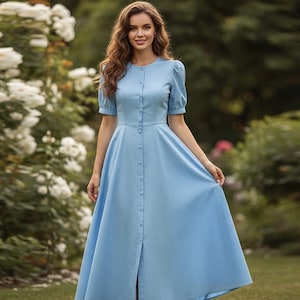 May include: A light blue, button-down dress with short, puffed sleeves. The dress has a fitted bodice and a flowing skirt. The model is standing in a garden with white flowers and green foliage in the background.