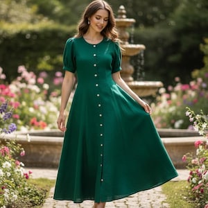 May include: A woman wearing a long, emerald green dress with short sleeves and a button-down front. The dress is flowing and elegant. The background features a garden with a fountain and colorful flowers.