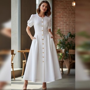 May include: A white button-down dress with short sleeves and a full skirt. The dress features gold-colored buttons and pockets. The model is wearing nude-colored heels. The background includes a brick wall and wooden furniture.