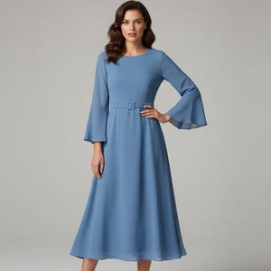 Pode incluir: Um vestido longo azul claro com mangas compridas e uma saia rodada. O vestido apresenta um pequeno cinto na cintura e mangas sino. A modelo est&aacute; vestindo o vestido em frente a um fundo cinza claro.