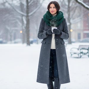 May include: A woman in a gray wool coat, green scarf, and black gloves holds a white mug in a snowy park. The coat is long and buttoned, and the scarf is a chunky knit. The background features snow-covered trees and a park bench.