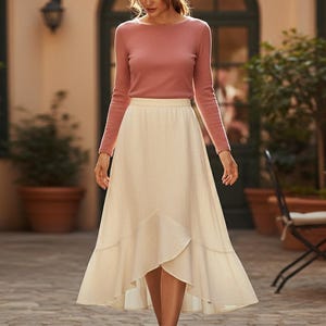 May include: A woman wearing a long-sleeved, dusty rose top and a cream-colored, high-waisted skirt. The skirt has a layered, asymmetrical hemline. The outfit is suitable for a casual or semi-formal occasion.