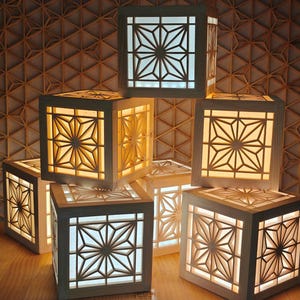 May include: Several illuminated wooden lanterns with detailed starburst designs. The cube-shaped lanterns emit a warm light. The background features a geometric wooden wall, creating a cohesive aesthetic.