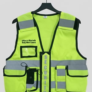 Op de afbeelding: Een felgele veiligheidsvest met zwarte randen, reflecterende zilveren strepen en meerdere zakken. Het vest heeft het woord "SECURITY" verticaal gedrukt en de tekst "Officer is fitted with Body Worn Camera".