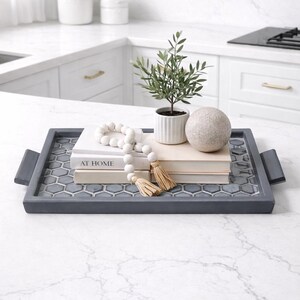 May include: A decorative tray with a gray frame and a mosaic tile base holds books, a small potted plant, a decorative sphere, and a beaded garland with tassels. The tray is on a white countertop.