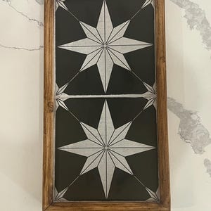 May include: A rectangular wooden serving tray with a dark background and a white starburst design. The tray has a light brown wooden frame and two wooden handles. The starburst design is repeated twice on the tray.