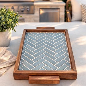 May include: A rectangular wooden serving tray with a blue herringbone tile design. The tray has a dark brown wooden frame and two wooden handles. The tray is sitting on a white surface, with a plant and outdoor kitchen in the background.