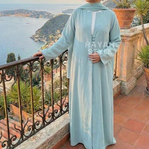 May include: A light blue long dress with long sleeves and a matching scarf. The dress has a button-up front and is worn outdoors. The text "HAYAH STYLE" is visible in the upper left and center of the dress.