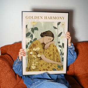 May include: A framed art print titled "GOLDEN HARMONY" from the "Pastoral Collection - 2022." The artwork depicts a woman in a yellow dress with a floral pattern, resting her head. The background includes white flowers and green foliage.