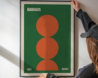 May include: A framed Bauhaus-style poster with a green background and three overlapping orange circles. The word "BAUHAUS" is at the top, with "1923" and "RETRO" at the bottom. The poster is held by a person.