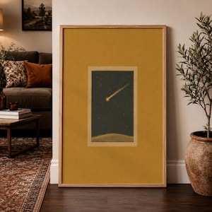 Op de afbeelding: Een rechthoekig ingelijst kunstwerk aan de muur met een mosterdgele passe-partout rondom een centrale illustratie van een gloeiende komeet die door een donkere nachtelijke hemel trekt boven een gebogen horizonlijn, in een licht houten lijst.