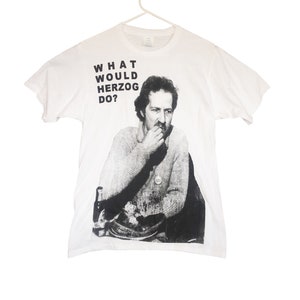 May include: White t-shirt with a black and white image of Werner Herzog, a famous filmmaker, with the text "WHAT WOULD HERZOG DO?" printed on the front.
