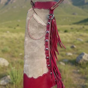Puede incluir: Un solo chaps de cuero marrón y rojo con flecos. Los chaps tienen un ribete rojo decorativo y un bolsillo. Los flecos son de color rojo intenso. Los chaps están diseñados para usarse sobre pantalones.
