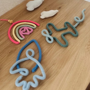 Op de afbeelding: Een verzameling handgemaakte decoraties voor de kinderkamer, waaronder een regenboog, een raket, wolken en abstracte vormen. De regenboog heeft roze, gele en groene kleuren. De raket is blauw. De wolken zijn wit. De vormen zijn blauw en groen.