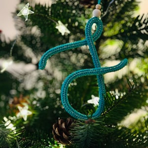 Könnte beinhalten: Türkisfarbener, gehäkelter Weihnachtsschmuck in Form eines stilisierten Buchstabens "J", der an einem Weihnachtsbaum hängt. Der Schmuck besteht aus dickem, strukturiertem Garn und wird von einer dünnen, hellfarbigen Schnur mit einer Holzperle gehalten.