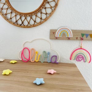 Puede incluir: Un letrero personalizado con el nombre Olivia en hilo rosa, naranja, amarillo, verde, azul y morado, con forma de nube. Incluye un perchero de madera con un gráfico de arcoíris, un arcoíris de hilo colgante y varias estrellas de fieltro.