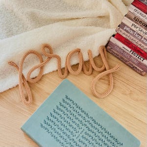 Könnte beinhalten: Ein hellbraunes, gestricktes Wortschild, das das Wort pray in Schreibschrift bildet. Das Schild befindet sich neben einem Stapel Bücher mit Titeln wie encourage, speak, persevere, practice, be patient, serve, protect sowie honor and respect.