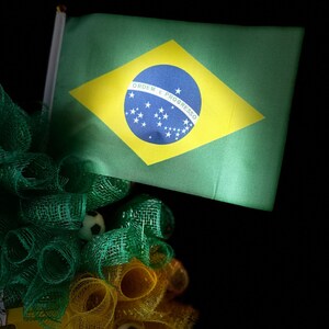 Puede incluir: Una bandera brasile&ntilde;a verde con un rombo amarillo y un c&iacute;rculo azul, con estrellas blancas y texto. La bandera forma parte de un arreglo decorativo con malla verde y amarilla y peque&ntilde;os balones de f&uacute;tbol, sobre un fondo negro.