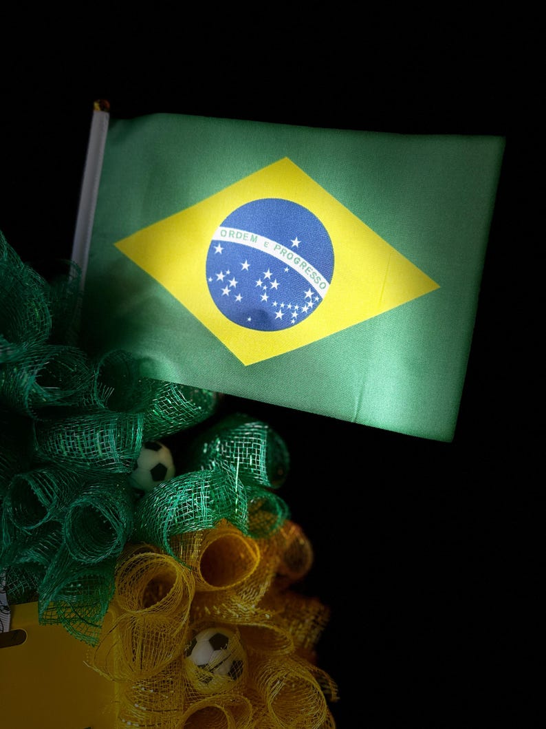 Puede incluir: Una bandera brasile&ntilde;a verde con un rombo amarillo y un c&iacute;rculo azul, con estrellas blancas y texto. La bandera forma parte de un arreglo decorativo con malla verde y amarilla y peque&ntilde;os balones de f&uacute;tbol, sobre un fondo negro.