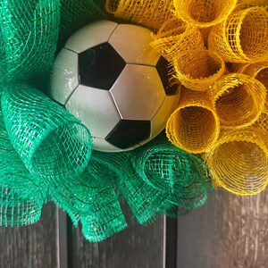 Puede incluir: Una corona decorativa con un bal&oacute;n de f&uacute;tbol en el centro, rodeada de cinta de malla verde y amarilla. El bal&oacute;n de f&uacute;tbol es blanco con pent&aacute;gonos negros. La cinta de malla est&aacute; dispuesta en un patr&oacute;n circular.