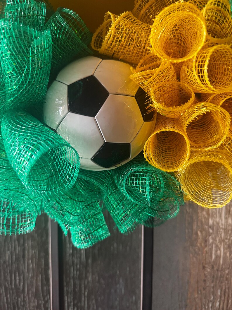 Puede incluir: Una corona decorativa con un bal&oacute;n de f&uacute;tbol en el centro, rodeada de cinta de malla verde y amarilla. El bal&oacute;n de f&uacute;tbol es blanco con pent&aacute;gonos negros. La cinta de malla est&aacute; dispuesta en un patr&oacute;n circular.