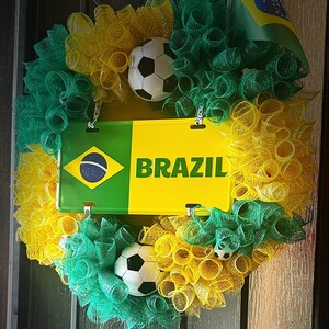 Puede incluir: Una corona decorativa con los colores de la bandera brasile&ntilde;a, con malla verde y amarilla, balones de f&uacute;tbol y una placa que dice "BRAZIL". Una peque&ntilde;a bandera brasile&ntilde;a est&aacute; adjunta.