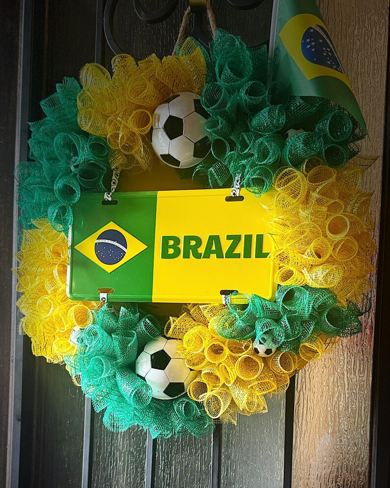 Puede incluir: Una corona decorativa con los colores de la bandera brasile&ntilde;a, con malla verde y amarilla, balones de f&uacute;tbol y una placa que dice "BRAZIL". Una peque&ntilde;a bandera brasile&ntilde;a est&aacute; adjunta.