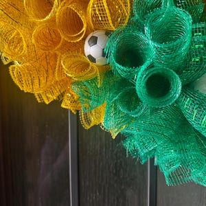 Puede incluir: Una corona decorativa con malla amarilla y verde, con un peque&ntilde;o bal&oacute;n de f&uacute;tbol en el centro. La corona est&aacute; dise&ntilde;ada con malla enrollada y es adecuada para la decoraci&oacute;n tem&aacute;tica deportiva.