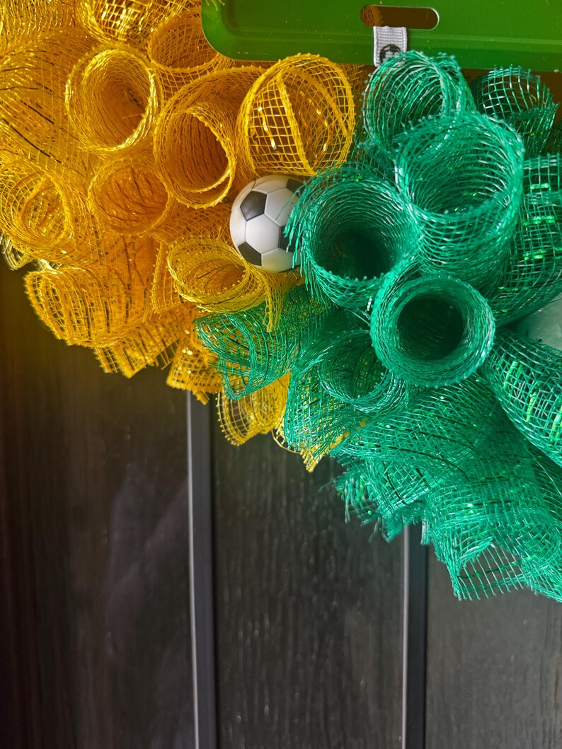Puede incluir: Una corona decorativa con malla amarilla y verde, con un peque&ntilde;o bal&oacute;n de f&uacute;tbol en el centro. La corona est&aacute; dise&ntilde;ada con malla enrollada y es adecuada para la decoraci&oacute;n tem&aacute;tica deportiva.