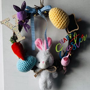 Puede incluir: Una corona de Pascua con un conejito de peluche blanco, un huevo tejido amarillo y un corazón tejido azul. La corona incluye una zanahoria roja, una cabeza de conejo morada y un letrero que dice "Happy Easter". La corona está decorada con una cinta de cuadros azul y blanca.