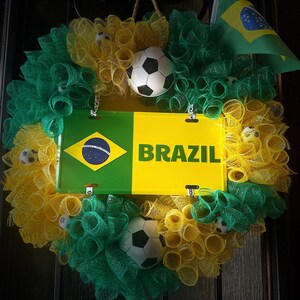 Puede incluir: Una corona decorativa con los colores de la bandera brasile&ntilde;a, con detalles en verde, amarillo y azul. La corona incluye un letrero que dice "BRAZIL" y una peque&ntilde;a bandera brasile&ntilde;a. Tambi&eacute;n se incorporan balones de f&uacute;tbol.