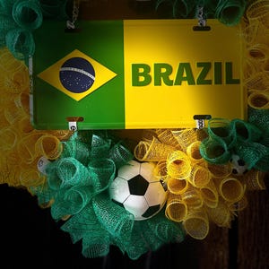 Puede incluir: Una corona decorativa con los colores de la bandera brasile&ntilde;a, verde y amarillo, con una placa que dice "BRAZIL". La corona incluye balones de f&uacute;tbol y cinta de malla.