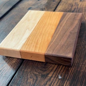 Puede incluir: Una tabla de cortar rectangular de madera con tres secciones de tonos de madera variables: marrón claro, medio y oscuro. La tabla está hecha de madera maciza y tiene una superficie lisa y acabada. La veta de la madera es visible.
