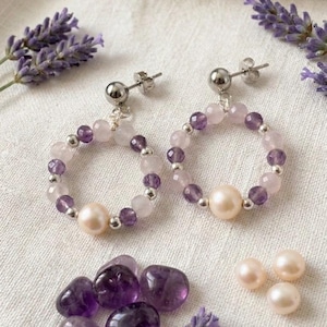 May include: A pair of hoop earrings with a stud backing, crafted with a combination of amethyst, rose quartz, and pearl beads. The earrings are accented with silver-colored metal beads. Additional amethyst stones and pearls are scattered nearby, with sprigs of lavender.