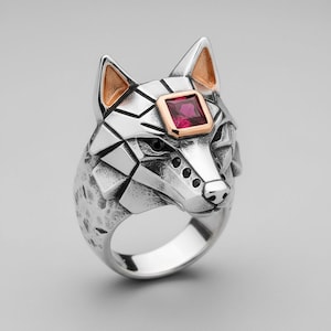 May include: A silver ring featuring a wolf head design. The wolf's head has geometric facets, with gold-colored accents on the ears and a square, red gemstone set in the forehead. The ring has a textured finish.