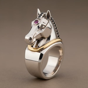 Puede incluir: Un anillo plateado con un diseño de cabeza de caballo. El caballo tiene una banda dorada alrededor del cuello y una pequeña gema roja redonda en la frente. La crin del caballo está detallada con detalles negros.
