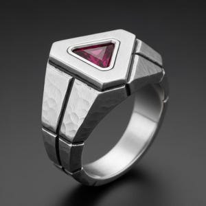 May include: A silver-toned ring with a geometric design. The ring features a triangular, ruby-colored gemstone set in a textured, angular frame. The band has a hammered texture, adding to its unique appearance.