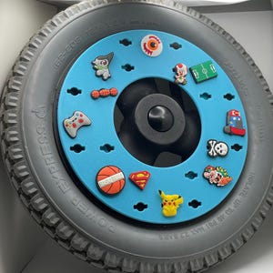May include: A gray wheelchair tire with a light blue center adorned with colorful charms. Charms include a basketball, a video game controller, and various cartoon characters. The tire is marked "POWER EXPRESS".