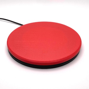 Op de afbeelding: Een rond, rood elektronisch apparaat met een zwarte basis en een zwart snoer. Het apparaat heeft een gestructureerd oppervlak en wordt waarschijnlijk gebruikt voor het bedienen van een computer of andere elektronische apparatuur.