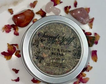 May include: A silver tin of "Sacred Spliff Herbals Focus & Flow Blend" (2 oz) surrounded by rose petals and gemstones. The tin has a clear top, displaying dried herbs. Red, pink, and clear gemstones are scattered around the tin.