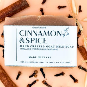 Puede incluir: Una barra de jabón artesanal de leche de cabra "Cinnamon & Spice" con palitos de canela, clavos y espigas de trigo. La etiqueta dice "Made in Texas" y pesa 113g. El jabón está sobre una superficie de color melocotón.