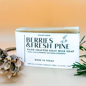 May include: A rectangular bar of goat milk soap in a white box. The box reads "BERRIES & FRESH PINE" and "HAND CRAFTED GOAT MILK SOAP". The soap weighs 113g. The background is a light orange colour.
