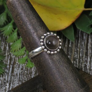 K&ouml;nnte beinhalten: Ein silberner Ring mit einem runden, klaren, rauchigen Quarzstein, der in einer Perlenfassung sitzt. Der Ring befindet sich auf einem h&ouml;lzernen Ringst&auml;nder.
