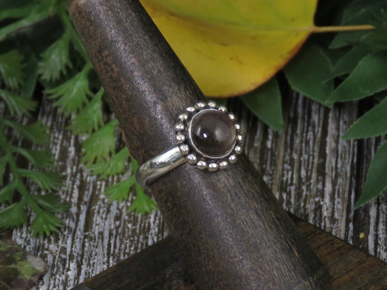 K&ouml;nnte beinhalten: Ein silberner Ring mit einem runden, klaren, rauchigen Quarzstein, der in einer Perlenfassung sitzt. Der Ring befindet sich auf einem h&ouml;lzernen Ringst&auml;nder.